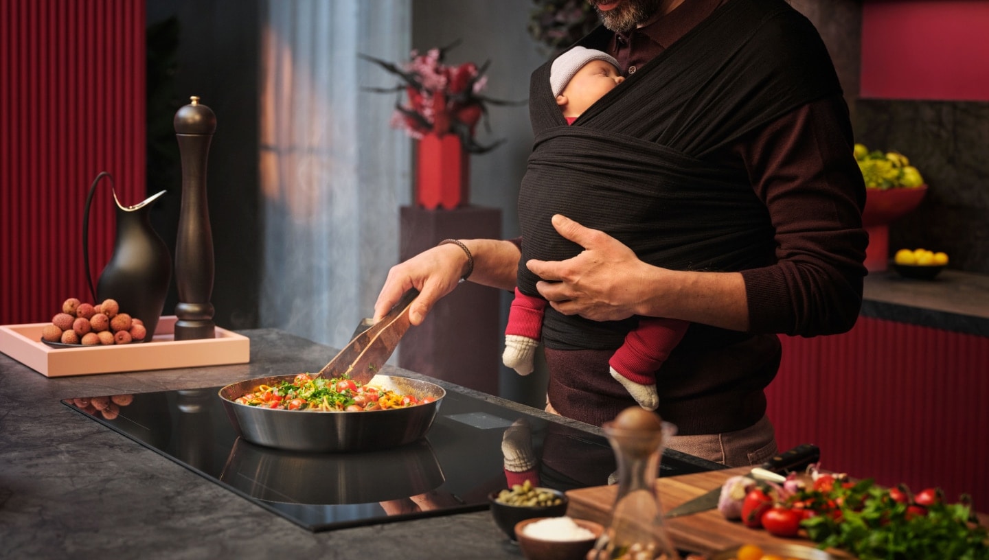 Man cooking food on a hob while carrying a baby in a baby carrier.